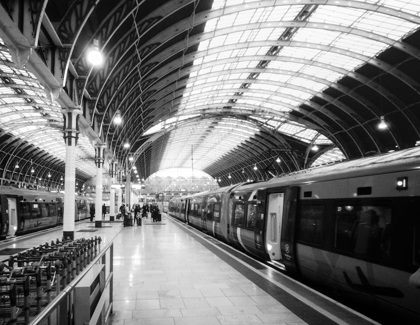 Inside a large train station with high, arched glass and metal roofing, a modern passenger train with dark tinted windows is stationary on the platform. Several people are visible in the background, some carrying luggage, near small groups of cardboard boxes and packing materials placed on the platform. The boxes are in various sizes, with some wrapped in plastic, and are likely part of a home relocation or moving process facilitated by Man with Van Paddington. The platform is well-lit with overhead lighting, and a trolley is positioned nearby, ready for loading or unloading. The scene captures the interior environment during a furniture transport or packing and moving operation, highlighting the logistics involved in house removals within an urban transport hub, with visible stairs and structural details supporting the station's architecture. The image demonstrates the movement and transfer of household items from the property to a transportation vehicle, emphasizing the process of packing and moving in a professional relocation context.