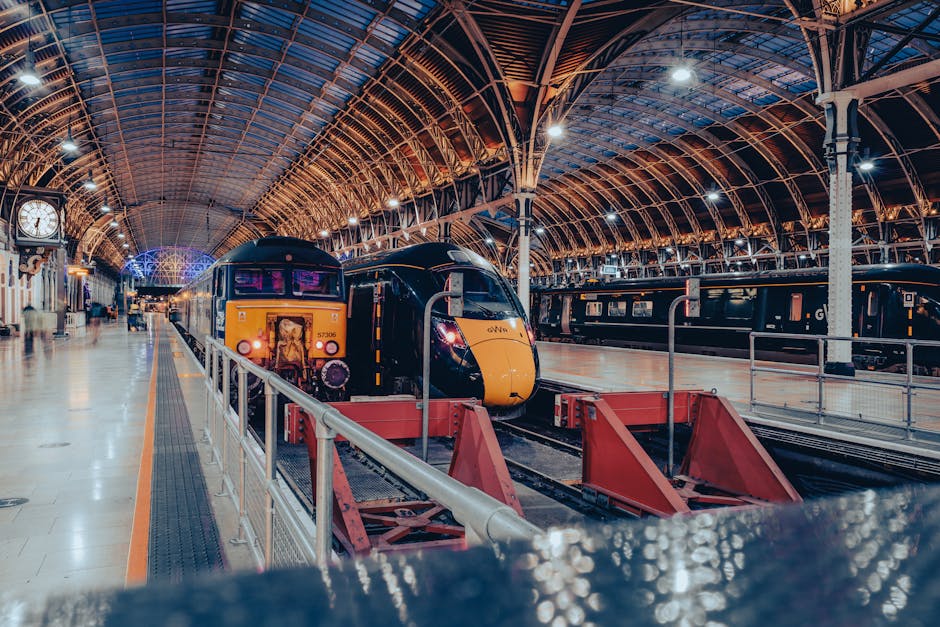 Inside a spacious train station with a high, arched metal and glass ceiling illuminated by bright white lights. A modern black and yellow passenger train is stationary on the platform, with a front view showing the train's headlights and a digital display screen. Adjacent to the train, there are platform barriers and a red cargo trolley with a flat surface used for loading or unloading luggage or large boxes. In the background, additional train carriages are visible, along with a large analog clock mounted on the wall near the platform edge, and a few passengers walking on the platform. The overall scene captures the environment of home relocation or packing and moving operations within a train station setting, with the train ready for departure or arrival as part of the moving logistics process, exemplifying professional removals and transport services.