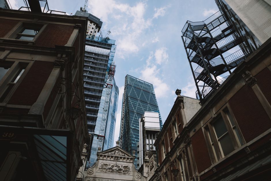 An upward view of a city street showing a mix of older and modern buildings, including a historic stone building with decorative carvings and a triangular pediment in the foreground, surrounded by contemporary glass skyscrapers under a partly cloudy sky. The older buildings display brick facades and traditional window styles, while the high-rise structures feature reflective glass exteriors and steel frameworks. A fire escape staircase is visible attached to one of the older buildings, and construction cranes can be seen atop some modern towers, indicating ongoing development. The scene captures the contrast and proximity of different architectural styles typical of urban environments like Paddington, with natural daylight emphasizing the textures of the building materials. This setting might be relevant for house removals, as it illustrates typical urban environments where relocation services provided by companies like Man with Van Paddington facilitate furniture transport, packing, and moving logistics in busy city streets.