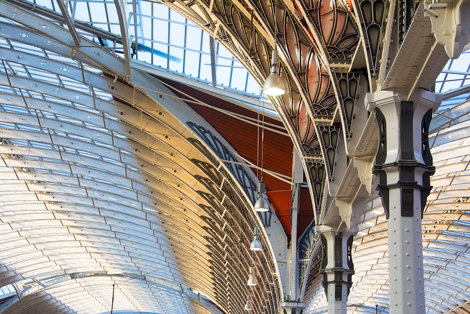 Interior view of a high-ceilinged building with ornate metal architectural supports painted black and white, supporting a curved glass roof structure with metal framing. Bright natural light filters through the glass panels, illuminating the intricate details of the structural elements. Several hanging pendant lights are suspended from the metal framework. In the foreground, there are no visible furniture or moving items, as this image primarily captures the architectural features of the indoor space, which could be part of a historical building or a station often involved in home relocation or moving logistics. The elegant design and spacious interior are typical of buildings where careful planning is necessary for furniture transport and packing during a house removal. Man with Van Paddington occasionally handles such moving projects, which may involve navigating similar architectural environments during loading or unloading processes.