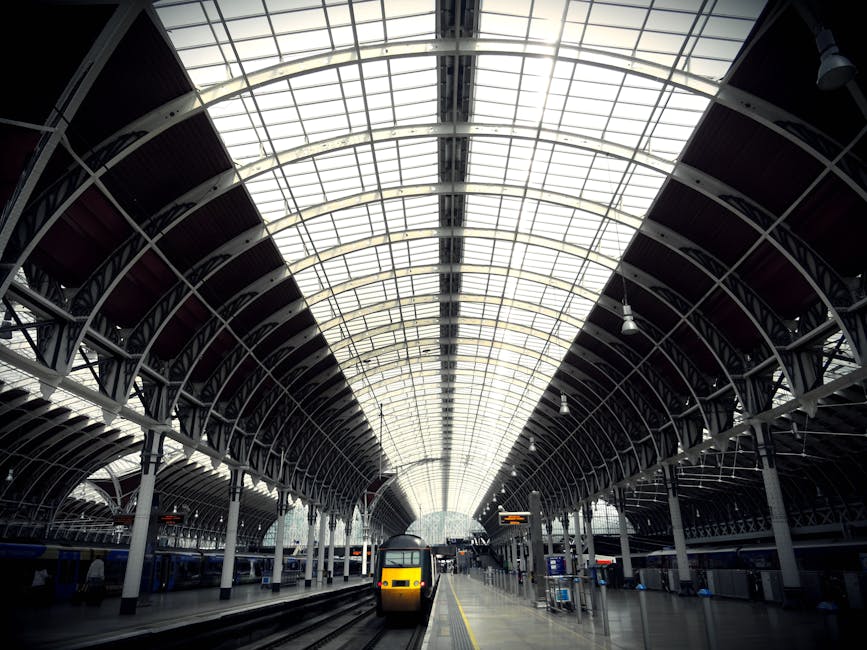 Inside a modern train station with a large, curved glass and metal roof structure supported by white columns. The platform is visible with a yellow and black train approaching or departing, and some luggage or packages may be seen nearby. The environment is well-lit with natural light filtering through the roof panels, and the scene captures the movement and logistical aspect of home relocation or furniture transport, emphasizing the importance of organized packing and loading processes in professional removals services like those provided by Man with Van Paddington. This setting illustrates the typical loading zone for moving household items into a vehicle, situated close to the station platform, highlighting the logistics involved in house removals within urban environments.