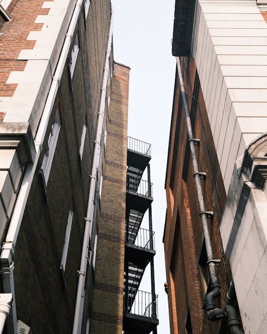 A view looking up between two narrow, multi-storey residential buildings in Paddington, with visible fire escape staircases on the exterior facades made of metal platforms and railings. The buildings feature brick and stone exteriors, with windows on each floor. The sky above is partly visible, indicating a tight urban street environment typical for house removals and moving services in areas with narrow streets and stairs. This setting highlights the challenges of furniture transport and loading during home relocation in Paddington, with the compact space emphasizing the importance of expert packing, careful maneuvering, and professional removals solutions provided by Man with Van Paddington.
