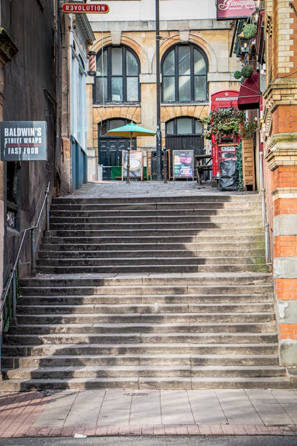 A series of wide, concrete steps leading up to a building entrance in a narrow Paddington street, with black metal handrails on both sides. To the left, there is a sign for Baldwin's Street Wraps and Fast Food, attached to a brick wall, and to the right, a red postal box is situated next to a brick building with potted plants on a small balcony. At the top of the stairs, the entrance features large arched windows and doors, with a black railing and a small outdoor area visible beyond. The scene suggests a typical urban setting suitable for house removals or furniture transport, with a focus on the layout and environment that Man with Van Paddington might encounter in home relocation or moving logistics services.