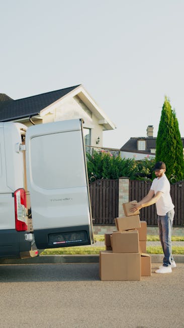 A man with a beard, wearing a white t-shirt, blue jeans, white sneakers, and a black cap, is sitting on the pavement at the rear of a black van during a home relocation process. The open van trunk is 