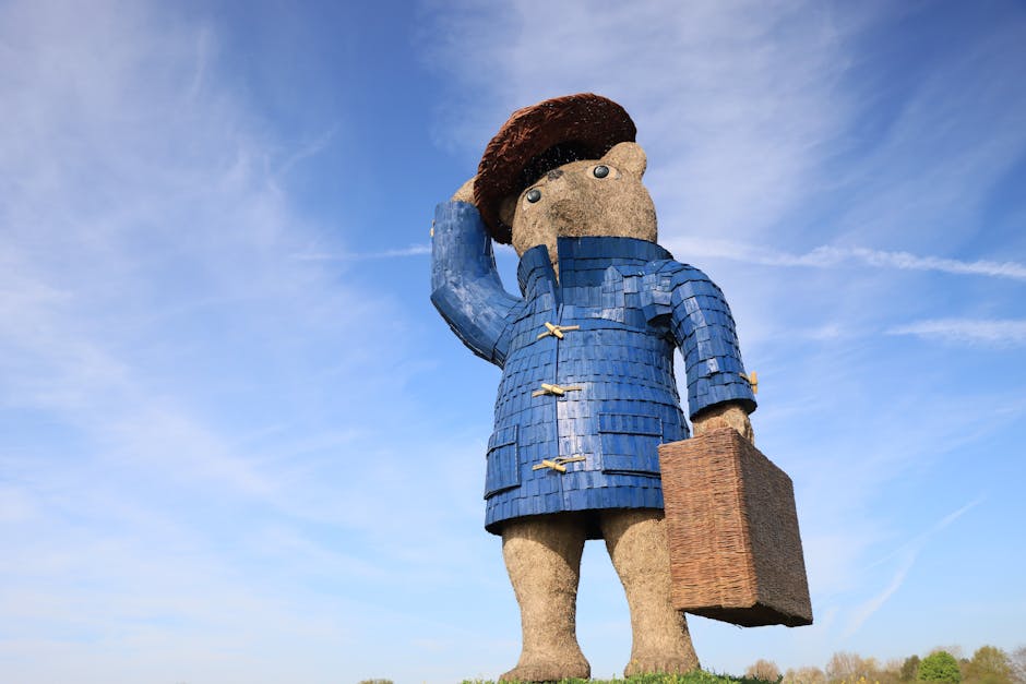 A large outdoor statue of a person with a textured, tan face and brown hair, wearing a blue, scale-like coat with wooden toggle buttons, stands against a backdrop of a clear blue sky with a few wispy clouds. The statue appears to be holding its right hand to its head in a saluting or thinking gesture, with the arm bent at the elbow. The figure's eyes are wide and cartoonish, with grey irises. The statue's environment suggests it is situated in a public space, possibly near a building or park, and it is made of various materials including textured stone or concrete for the face and fabric-like or wooden textures for the clothing. The lighting is natural, indicating daytime, and the overall scene emphasizes the size and detailed craftsmanship of the statue, which could be related to cultural or artistic displays in urban or park settings. Occasionally, a company such as Man with Van Paddington might have scheduled furniture transport or moving services nearby, supporting home relocation or packing and moving processes.