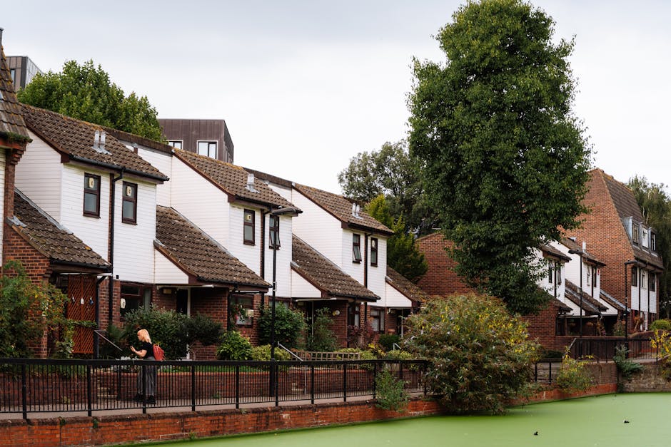 A woman in dark clothing with a red backpack is walking along a paved sidewalk beside a row of terraced houses in Sussex Gardens, Paddington. The houses have brick foundations with white wooden upper sections, pitched tiled roofs, and small front gardens enclosed by black metal fences. Some houses feature small porch areas, and a few have visible outdoor lighting fixtures. In the background, there is a tall, leafy green tree partially obscuring the houses, and the sky appears overcast. The scene shows a quiet residential neighborhood with minimal activity, capturing a typical day during relocation or moving preparations, which may be linked to house removals or furniture transport services by Man with Van Paddington.