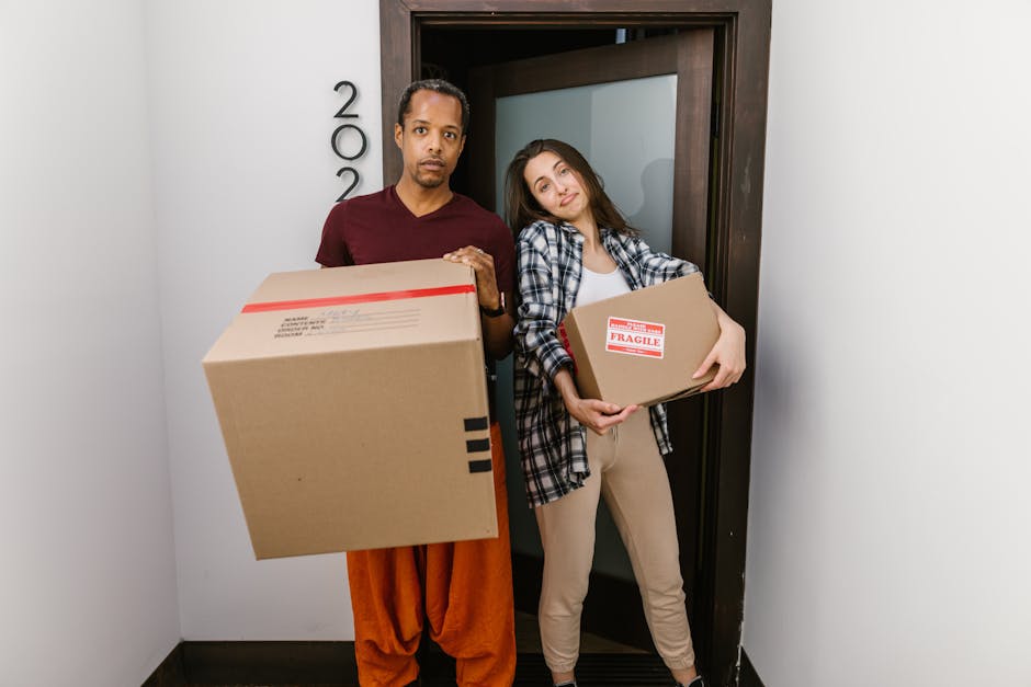 Two movers from Man with Van Paddington are positioned in a doorway during a home relocation process, each holding cardboard boxes. The mover on the left, wearing an orange outfit, holds a large box with red and black markings, while the mover on the right, dressed in casual beige pants and a plaid shirt, carries a smaller box labeled 'Fragile' in red text. The background shows an interior wall and a dark wooden door frame, suggesting they are inside an apartment or house. The scene captures the loading process involved in furniture transport and packing and moving services, with natural lighting illuminating the space, reflecting careful handling of household items as part of the house removals or relocation services offered by [COMPANY_NAME], aligned with the page's focus on trusted Paddington removals.