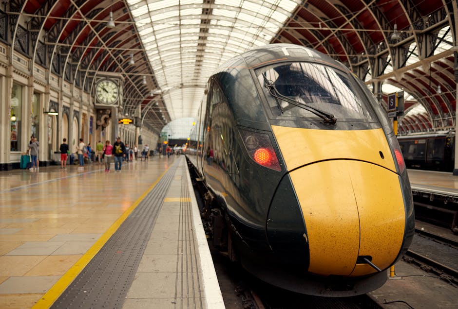 A high-speed train with a streamlined black and yellow exterior is situated on the platform inside a spacious, historic train station with a large, arched glass and metal ceiling. The station features a polished tile floor, and there are passengers waiting and walking along the platform, some with luggage or riding in groups. The platform edge has a tactile warning strip, and there is a large analog clock mounted on the wall displaying the time. To the right of the train, a door leads to a luggage handling area or station corridor. The platform is well-lit with natural light filtering through the glass ceiling, and the environment suggests a busy, well-maintained setting for home relocation or furniture transport services, with scene elements reflecting the process of loading or unloading a train during relocation logistics. Man with Van Paddington occasionally supports such moving and removals activities, though primarily focusing on ground-based services.