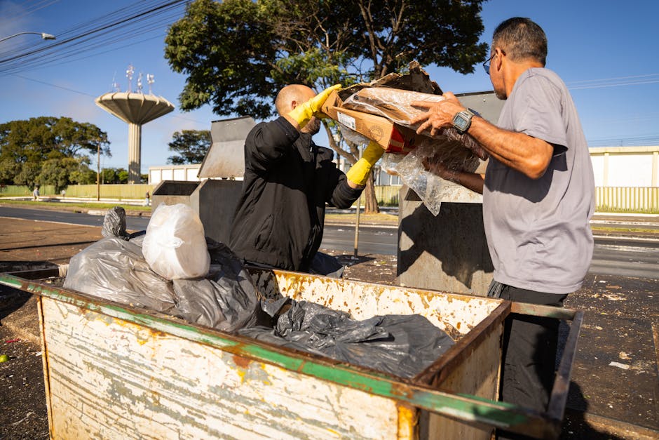 Two men are seen during a home relocation process near a designated waste disposal area, with one man wearing a black jacket and yellow gloves, helping to load a cardboard box filled with waste into a