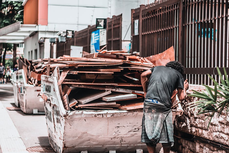Two men are seen during a home relocation process near a designated waste disposal area, with one man wearing a black jacket and yellow gloves, helping to load a cardboard box filled with waste into a